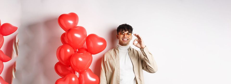 Valentines and romance concept. Happy young man smiling, showing OK sign near red hearts gesture, recommending good offer on lovers day.
