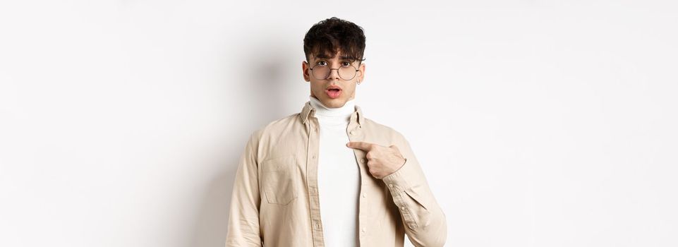 Confused and surprised young man pointing at himself, being chosen or accused, standing on white background.