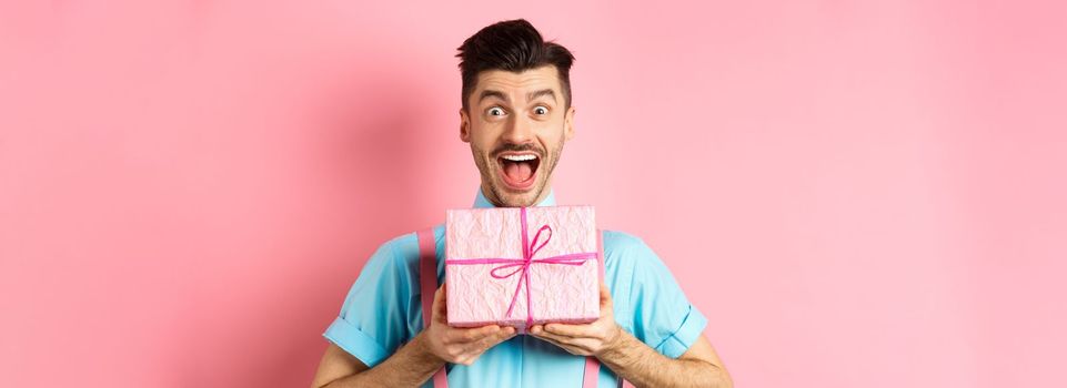 Holidays and celebration concept. Excited and surprised guy celebrating birthday, receiving gift and cheering, smiling happy at camera, standing over pink background.