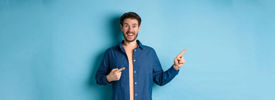 Excited and happy caucasian man smiling at camera, pointing fingers left at empty space, showing logo, standing on blue background. Copy space