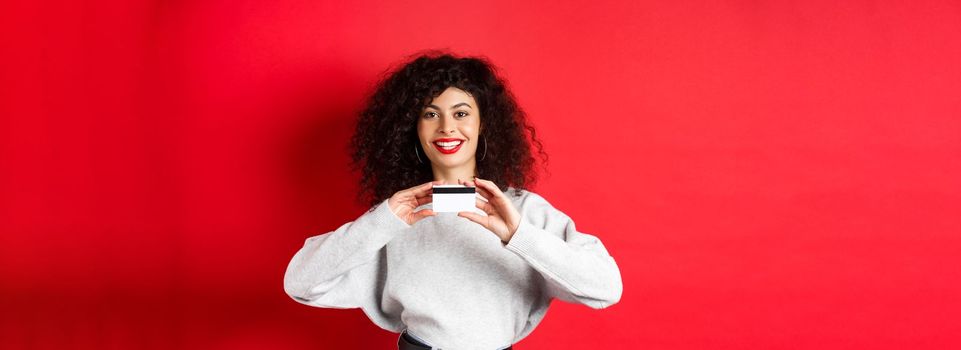 Happy lady with curly hair showing plastic credit card and smiling, standing against red background. Shopping concept.