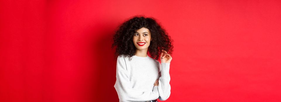 Modern woman in sweatshirt, playing with curly hair and looking aside at empty space, standing on red background.