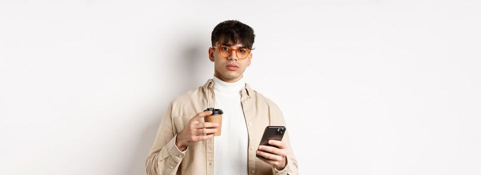 Real people. Stylish hipster guy in glasses holding cup of coffee from cafe takeaway and mobile phone, standing on white background.