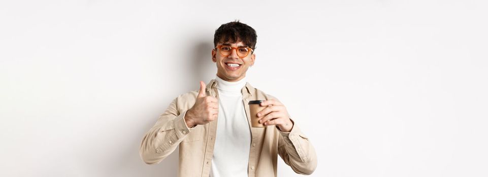 Real people. Cheerful smiling guy in glasses drinking coffee and showing thumb up, recommending cafe, standing on white background.