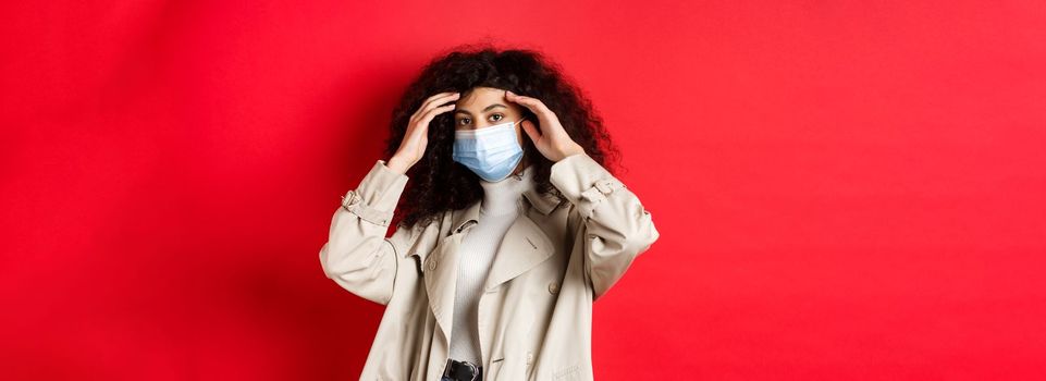 Covid-19, pandemic and quarantine concept. Stylish young woman with curly hair, going outside in medical mask and trench coat, fixing haircut, standing on red background.