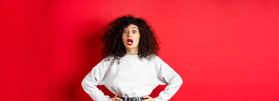 Shocked italian woman with curly hair, gasping and staring at camera astonished, open mouth, standing in white sweatshirt on red background.