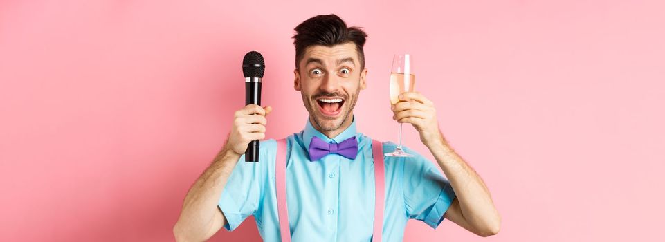 Party and festive events concept. Cheerful young male entertainer, giving speech on holiday, raising glass of chamapgne and holding microphone, making toast on wedding, pink background.