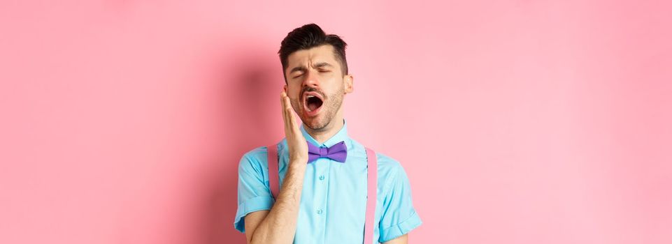 Image of tired guy in bow-tie yawning with opened mouth and closed eyes, standing sleepy, feeling fatigue, standing over pink background.