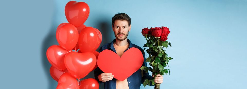 Valentines day romance. Young man with bouquet of red roses and heart balloons smiling, bring presents for lover on valentine date, standing over blue background.