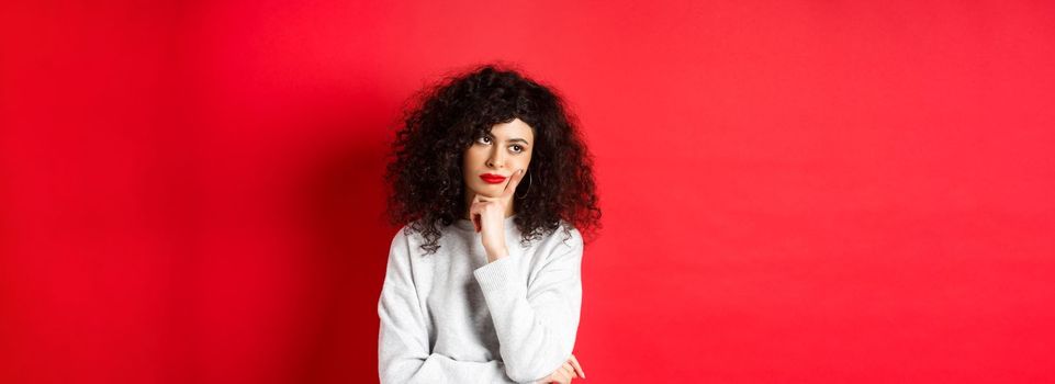 Grumpy young woman with curly hair, looking annoyed or bored at empty space, standing pensive and sad on red background.