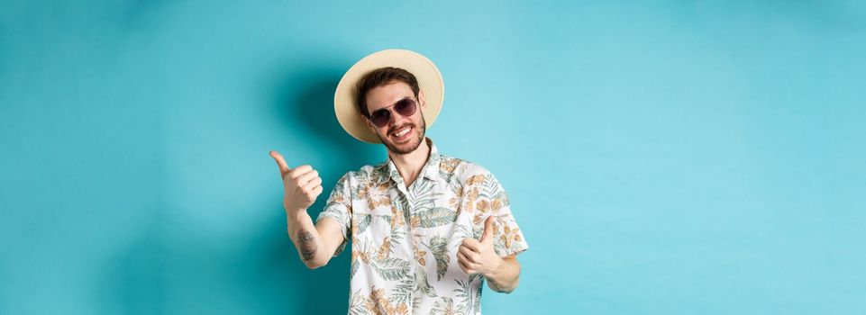 Cheerful tourist having fun summer holidays, showing thumbs up and smiling, standing in hawaiian shirt and sunglasses on blue background.