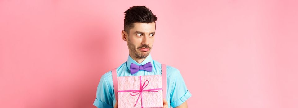 Holidays and celebration concept. Shocked guy staring aside with cringe face, holding small gift box, standing over pink background.