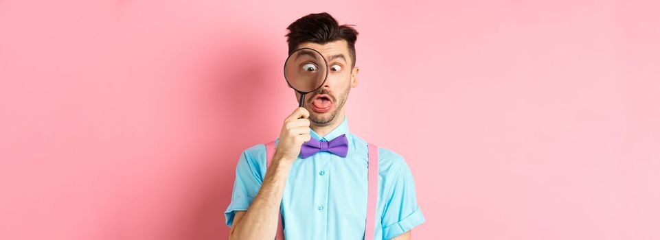 Funny man in bow-tie look through magnifying glass, squinting and making silly faces, standing on pink background.