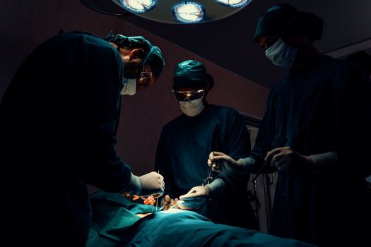 Surgical team performing surgery to patient in sterile operating room. In a surgery room lit by a lamp, a professional and confident surgical team provides medical care to an unconscious patient.