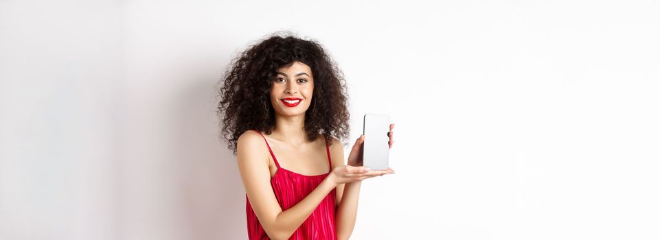 Elegant woman in red dress and makeup, showing blank smartphone screen and smiling, standing over white background. Copy space