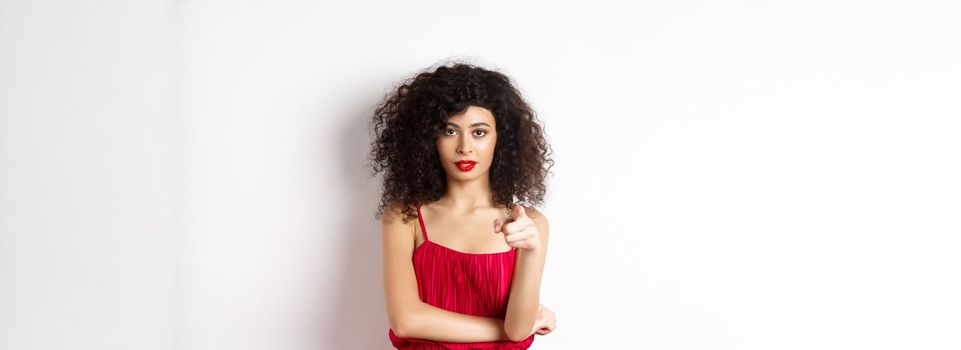 Confident young woman in red dress and makeup, pointing at camera, choosing you, inviting to event, standing over white background.