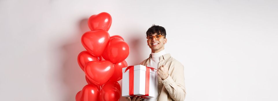 Valentines day and romance concept. Happy and confident boyfriend prepare gift for lover, saying yes and smiling, holding romantic gift, standing near red hearts balloons.