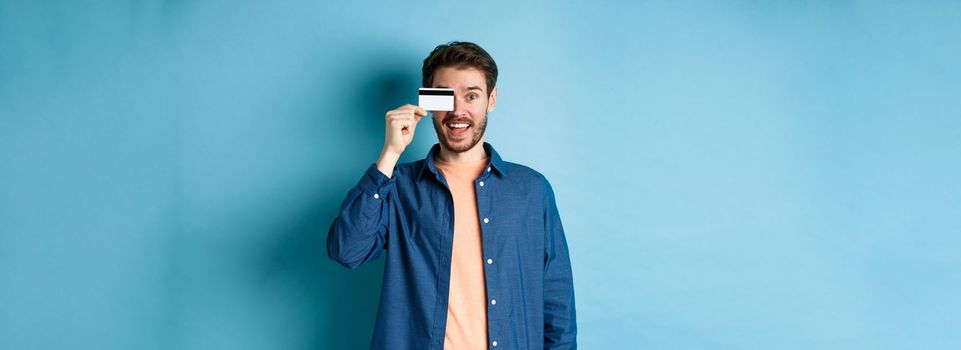 Cheerful handsome man showing plastic credit card and looking excited, checking out special offer, standing on blue background.