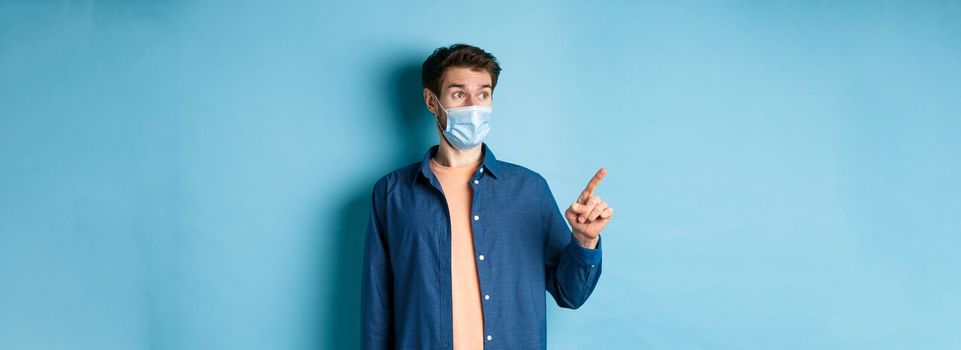 Covid-19 and healthcare concept. Surprised guy in face mask pointing and looking left at empty space, standing on blue background.