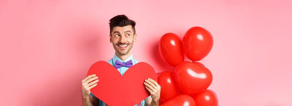 Valentines day concept. Romantic guy smiling and looking left, dreaming of date with lover, showing red big heart cutout, pink background.