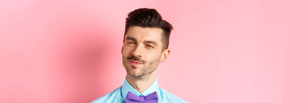 Close-up of smiling young guy with moustache, squinting intrigued, listening to something interesting, standing in bow-tie on pink background.