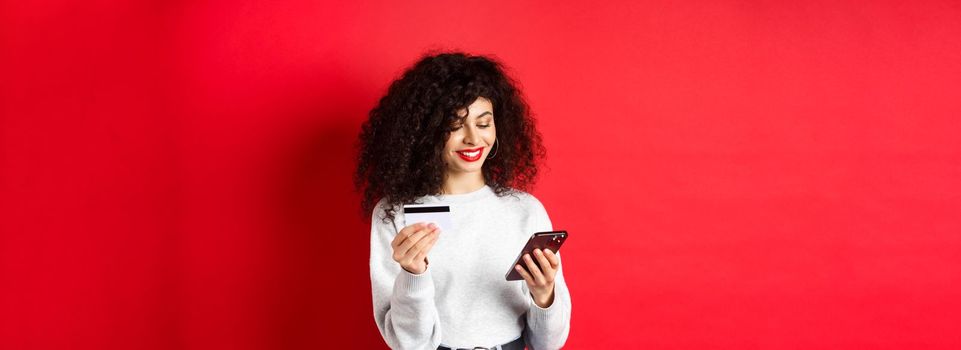 E-commerce and online shopping concept. Attractive caucasian woman paying for purchase in internet, holding smartphone and credit card, red background.