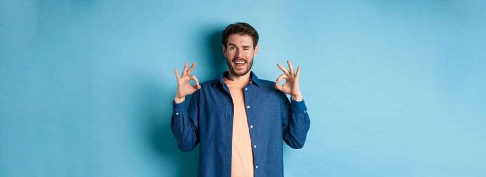 Happy smiling man winking and showing alright OK sign, say yes, approve and agree to something good, standing on blue background.