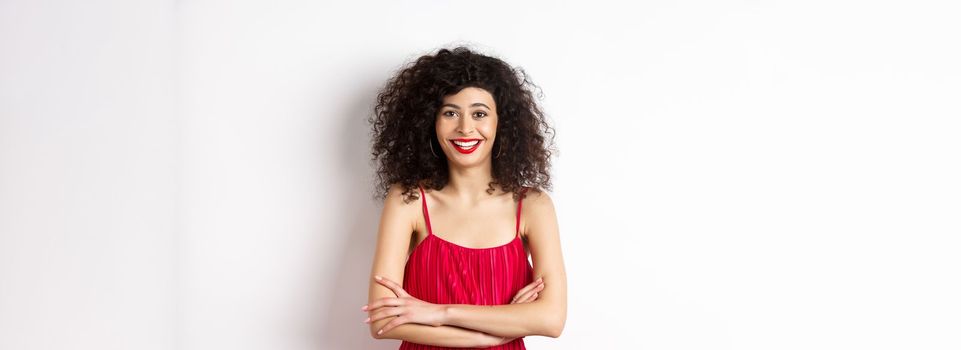 Elegant young woman in red dress with makeup, dressed-up for festive event, smiling happy at camera, standing over white background.