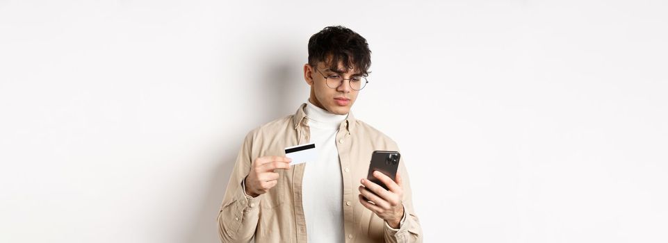 Handsome young man in glasses making purchase on phone, shopping online, holding plastic credit card and smartphone, standing on white background.