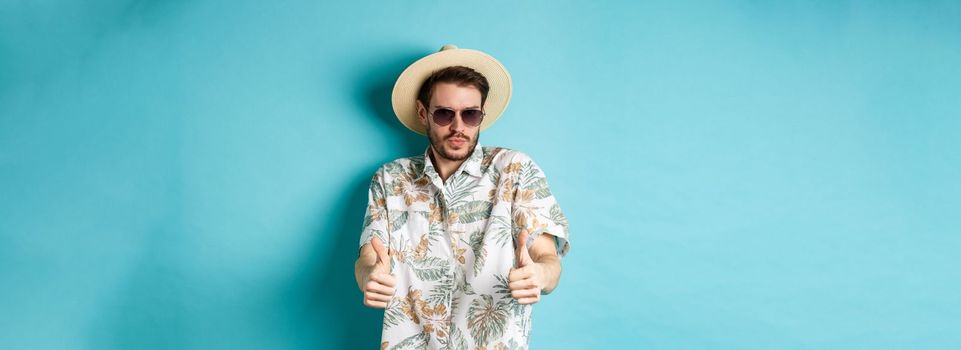 Handsome tourist enjoying summer holiday, wearing sunglasses and hawaiian shirt, showing thumbs-up in approval, standing on blue background.