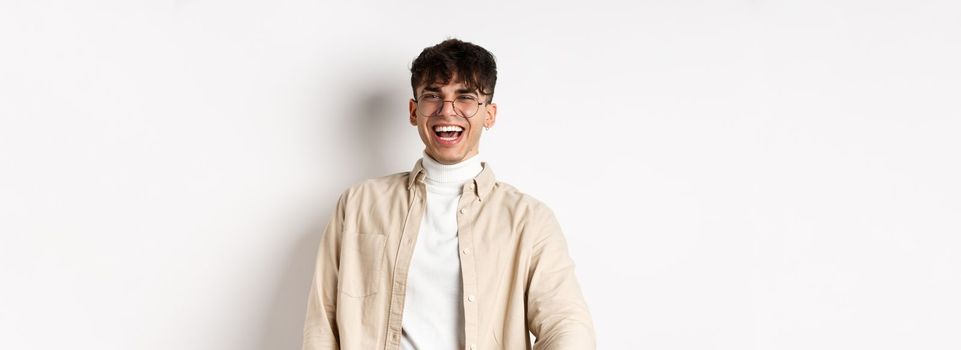 Image of happy young man laughing and looking upbeat, watching something funny, standing on white background. Authentic people concept
