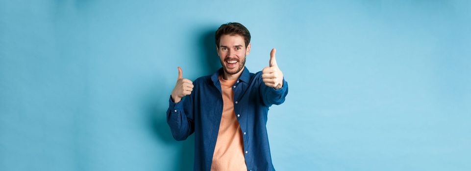 Positive smiling guy showing thumbs up and smiling, complimenting you, praising good job, well done gesture, standing on blue background.