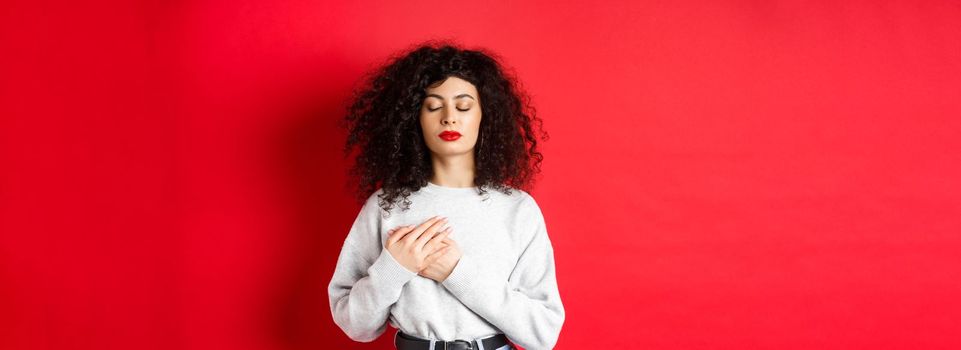 Image of calm young woman with curly hairsty, close eyes and holding hands on heart, keeping warm memories, feeling nostalgic, standing on red background.