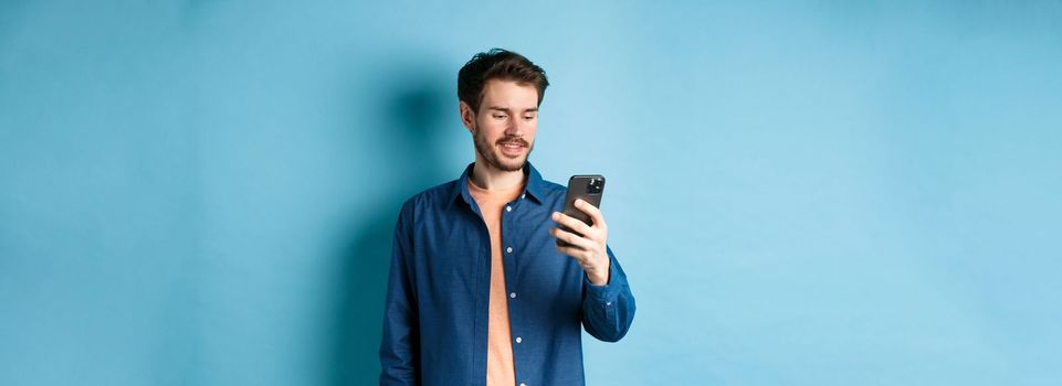 Handsome modern guy using mobile phone, reading smartphone screen and smiling, networking on blue background.