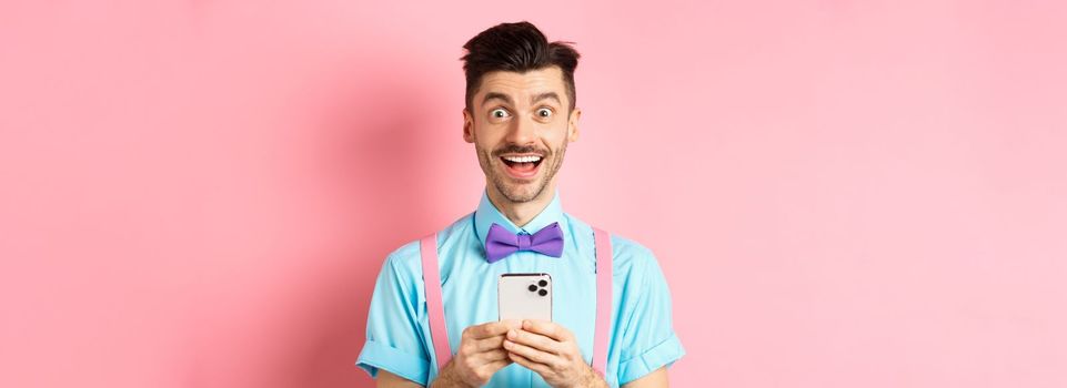 Online shopping. Happy man looking amazed after reading smartphone screen, smiling excited at camera, standing over pink background.