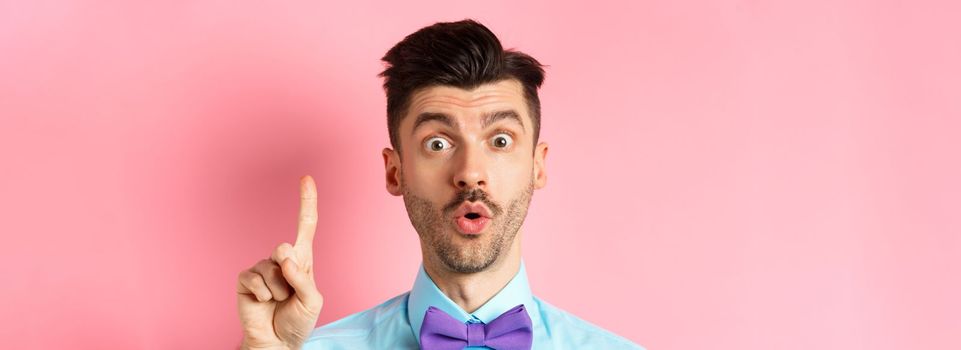 Close-up of excited funny guy with moustache and bow-tie, pitching an idea, raising finger to say suggestion, have plan, standing over pink background.