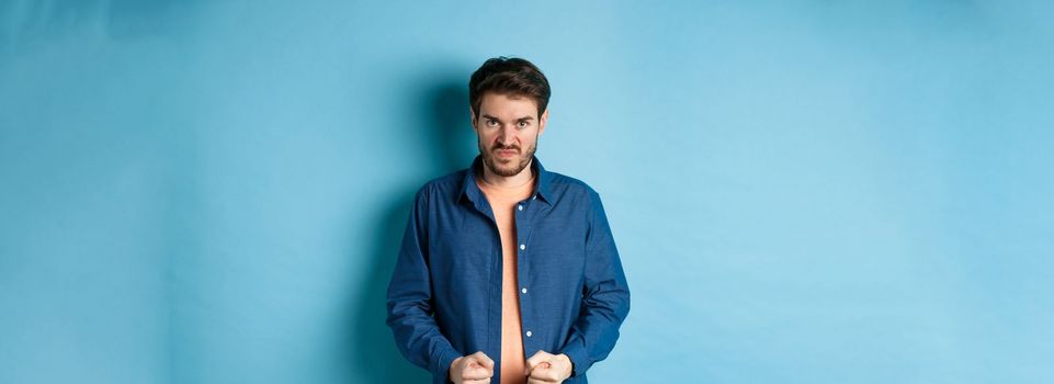 Angry and pissed-off man clench fists and stare with disdain at camera, trying to keep himself together, look with rage, standing on blue background.