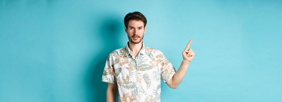 Portrait of handsome bearded guy on vacation, wearing summer shirt and pointing finger left at logo, standing on blue background.