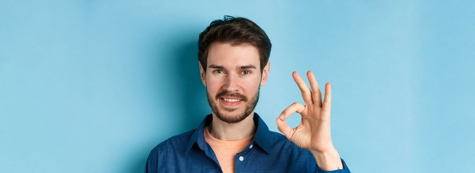 Close up of handsome bearded guy showing okay gesture and smiling, standing on blue background.