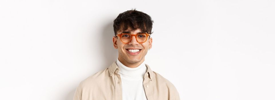 Close-up of handsome hipster guy with natural smile, wearing glasses and earrings, looking happy at camera, standing on white background.