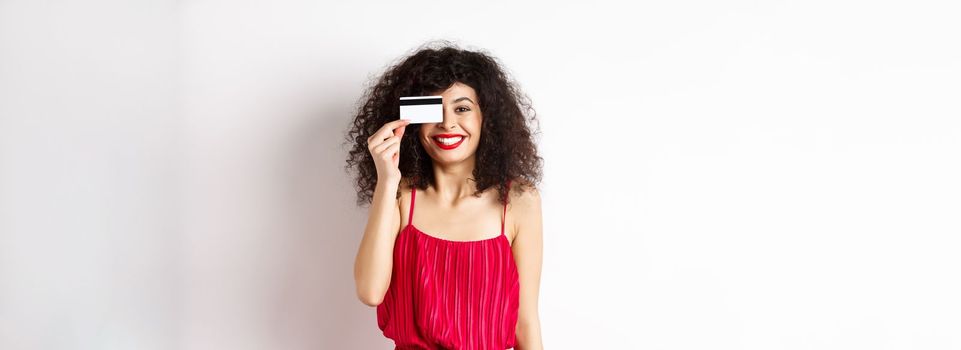 Image of elegant woman in red dress, showing plastic credit card and smiling at camera, standing over white background. Copy space
