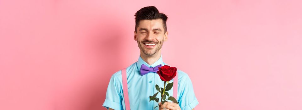 Valentines day and romance concept. Romantic man with red rose going on date with lover, standing in fancy bow-tie on pink background.
