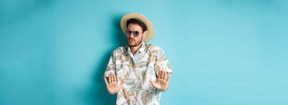 Alarmed tourist asking to stay away, step back from something cringe, showing rejection gesture, standing in straw hat and hawaiian shirt, blue background.