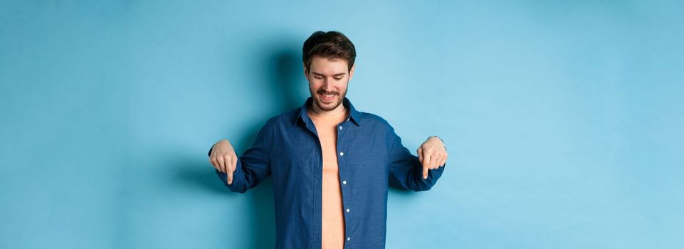 Smiling handsome man with beard, looking and pointing down at banner, checking out special offer, standing on blue background.