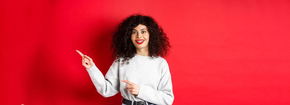 Amused young woman with curly hairstyle, pointing fingers right at logo, looking impressed and excited, checking out promotion, red background.