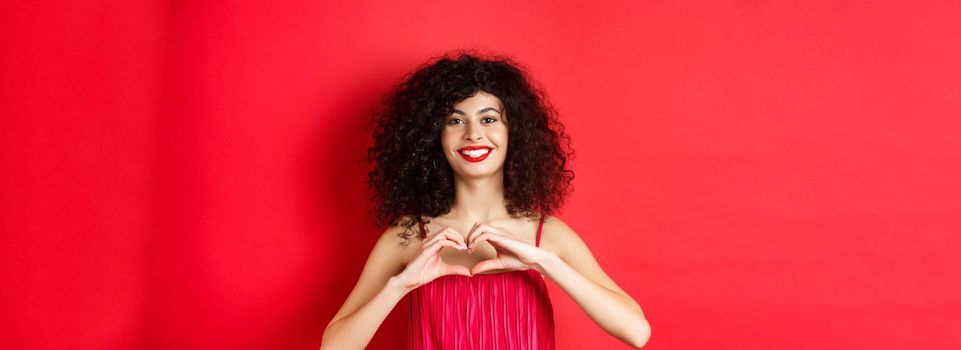 Valentines day. Romantic girl with curly hairstyle in evening dress, smiling and showing heart sign, say I love you on lovers holiday, standing over red background.