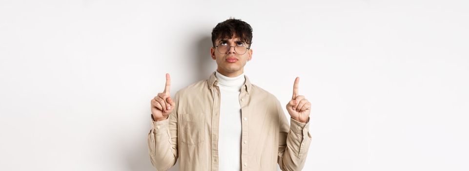 Image of annoyed or offended young man looking, pointing fingers up bothered, frowning disappointed, standing on white background.