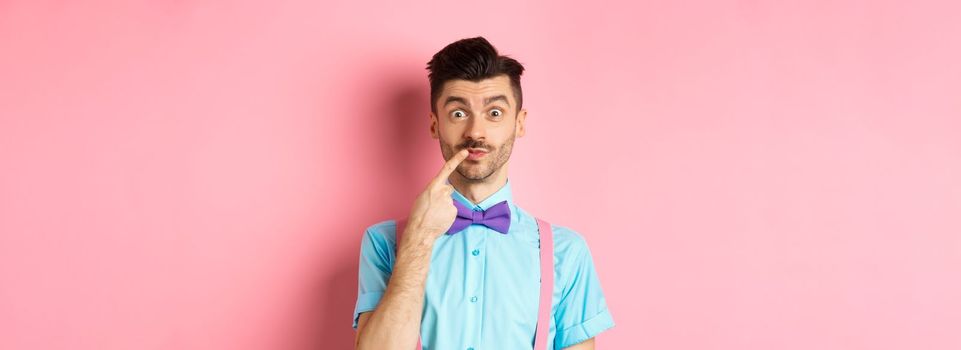 Intrigued happy guy touching lip, looking at camera pensive, making choice what to buy, standing over pink background.