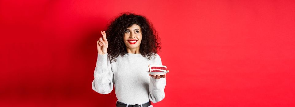 Celebration and holidays concept. Hopeful young woman making birthday wish, holding fingers crossed and looking up, holding b-day cake with candle, red background.