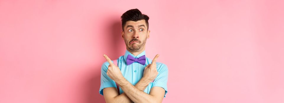 Puzzled funny man in bow-tie pointing fingers sideways, showing two variants on both sides, looking confused, standing over pink background.